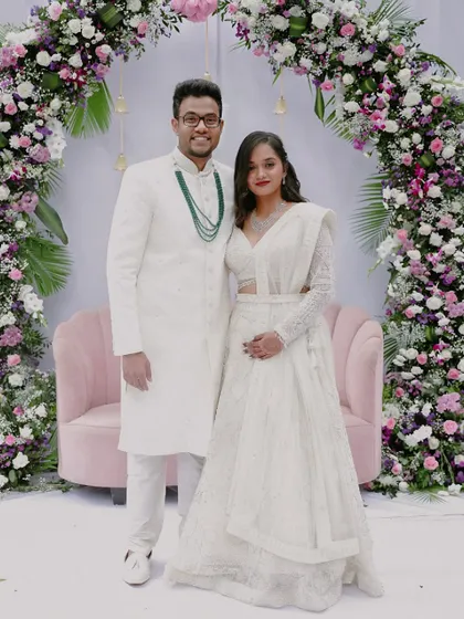 An elegant reception portrait of the couple in coordinated white outfits. Standing before a beautiful floral arch, this photo captures the classic, formal side of their wedding celebration.