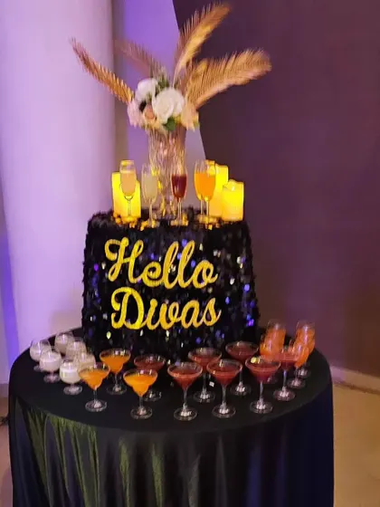 A 'Hello Divas' welcome drink station. The sequined table, feather arrangements, and a variety of colorful cocktails make for an inviting and glamorous start to the party.