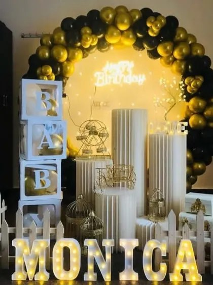 A chic black and gold birthday theme for Monica. The setup includes a balloon arch, 'BABY' blocks, a white picket fence, and various elegant props on display pedestals.
