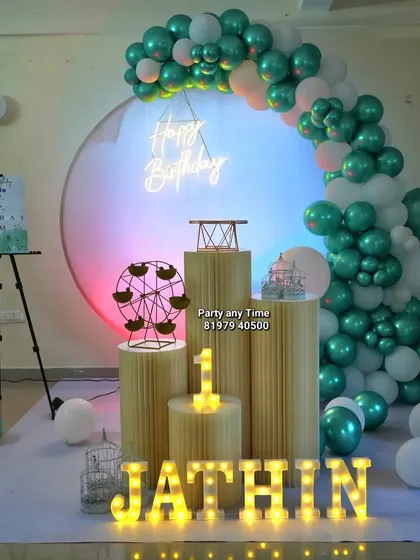 An elegant first birthday decoration with a teal and white balloon arch against a circular backdrop with a neon sign.