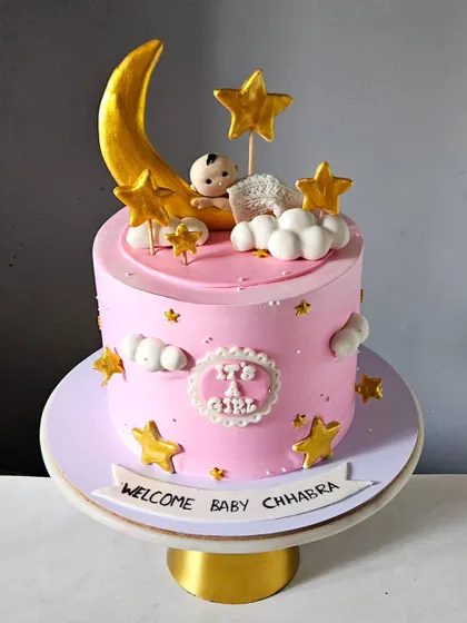 A sweet "Welcome Baby" cake for a baby girl. The pink cake is decorated with a golden moon, stars, and a tiny, sleeping baby topper, making it a perfect way to celebrate a new arrival.