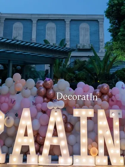 A close-up of the marquee letters for "RAATNA," framed by custom butterfly cutouts and a dense wall of pink, white, and rose gold balloons.