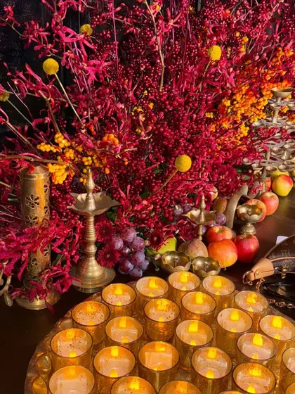 A close-up of a Diwali decor setup, with a tray of glowing candles, brass lamps, and vibrant red and yellow foliage.