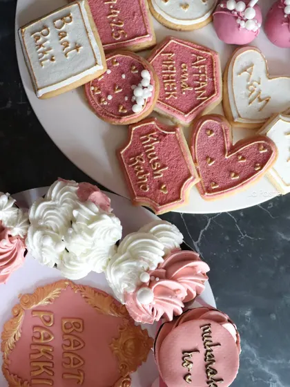 A beautiful spread of my engagement desserts, including custom-shaped cookies, cupcakes, and the central "Baat Pakki" plaque.