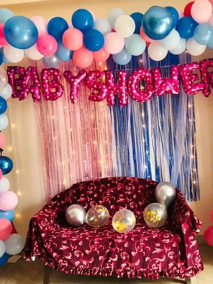 A home baby shower decoration with a pink and blue theme. The setup includes a foil curtain backdrop and a "BABY SHOWER" foil balloon banner.