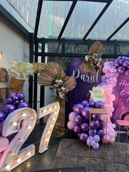 A chic and trendy decoration for a 27th birthday, using a rich purple marble-effect backdrop, metallic balloons, and dried palm leaf accents.