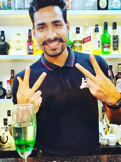 A happy student flashing a peace sign behind the bar, surrounded by a wide selection of spirits.
