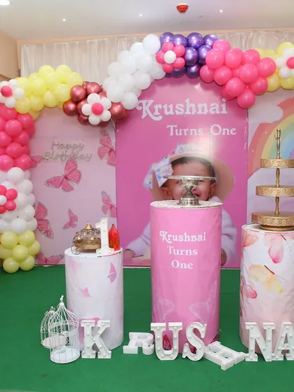 This butterfly-themed first birthday setup includes a personalized backdrop with the child's photo, a multi-colored balloon arch, and decorative plinths for the cake.