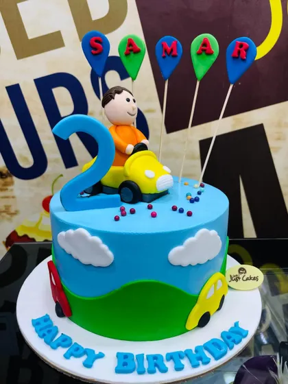A bright blue 2kg cake for a second birthday, topped with a boy in a car and balloon-themed name toppers.