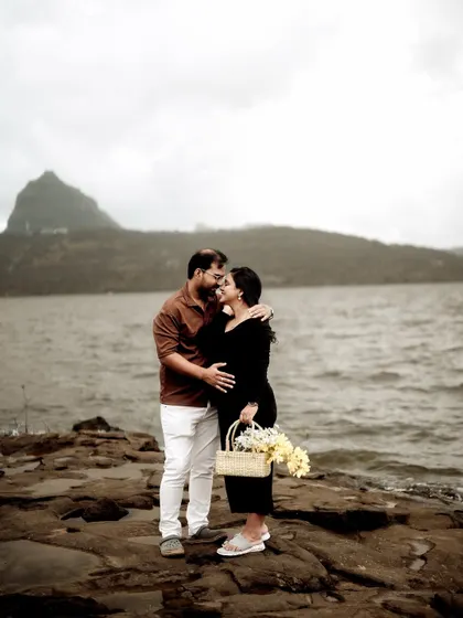 A romantic embrace on a rocky shore. He kisses her forehead as they stand by the water, a basket of flowers in her hand, creating a beautiful, storybook moment.
