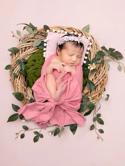 A beautifully composed overhead shot of a newborn wrapped in dusty pink, resting in a wicker wreath adorned with green leaves. The simplicity of the setup highlights the baby's peacefulness.