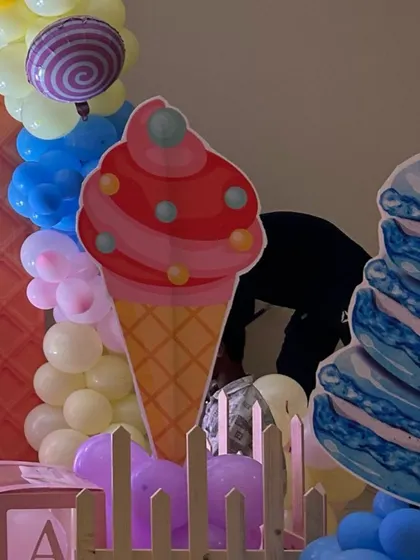 A close-up of a candyland party, showing a large ice cream cone cutout next to a stack of giant macarons and a colorful balloon garland.