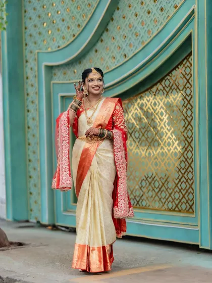 This full-length shot of the Telugu bride showcases the beautiful fall of the pre-pleated saree. The contrast of the off-white silk with the vibrant red border and blouse creates a striking and traditional look.