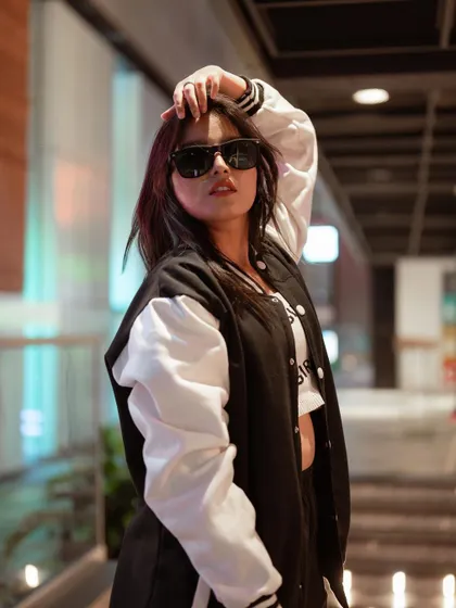 This portrait captures a cool, confident attitude. The model's varsity jacket and sunglasses, combined with the blurred urban background, create a classic street style look.