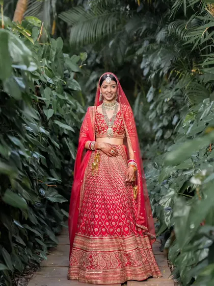 A full-length portrait of the bride in her red wedding lehenga, standing in a lush green pathway in Goa.