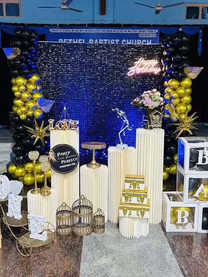 This large-scale birthday decoration features a stunning blue shimmer wall as the centerpiece. The wall is framed by black and gold balloon columns, multiple pedestals, and 'BABY' blocks, creating a grand display.