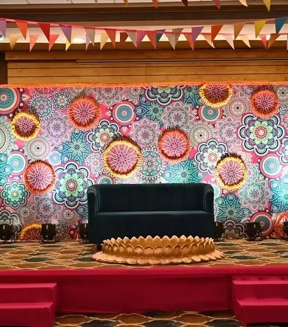 A vibrant and colorful stage backdrop for a Haldi function. The design uses a mandala-printed flex and decorative hangings to create a festive and energetic photo corner.