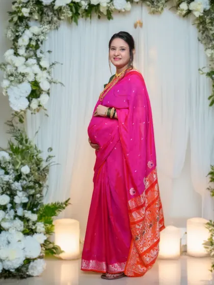 A standing portrait of an expecting mother in a bright pink saree, framed by a beautiful white floral arch in the studio. The soft lighting creates a warm and celebratory atmosphere.