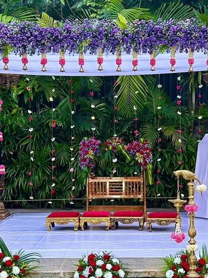 A traditional naming ceremony mandap with carved wooden pillars, wrapped in garlands of red and pink flowers, set against a lush green backdrop.