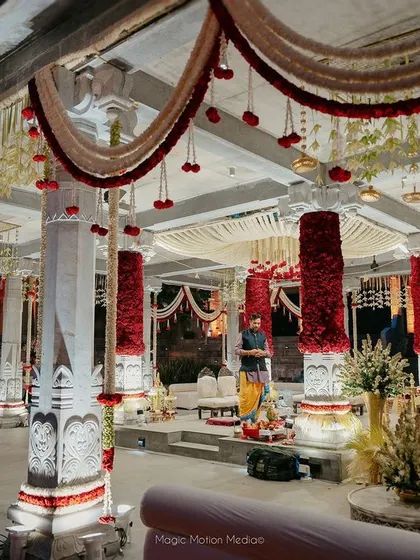 Inside the mandap, where pillars are wrapped in dense red roses and the ceiling is draped with garlands of jasmine and red flowers, creating a rich and fragrant canopy.