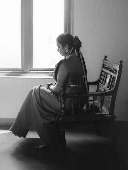 A black and white portrait of a bride seated on a wooden bench, looking out a window. The image is full of quiet contemplation and timeless grace.