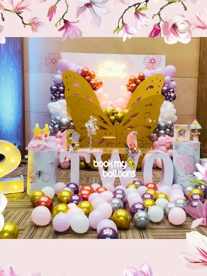 A whimsical butterfly-themed second birthday party featuring large golden butterfly wings as the central photo-op, surrounded by pink, purple, and gold balloons.