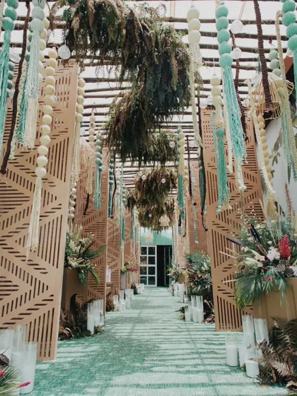 The entrance to a tropical-themed welcome party, featuring a walkway lined with laser-cut wooden panels and an overhead installation of macrame tassels and dried ferns, immediately immersing guests in a chic, bohemian vibe.