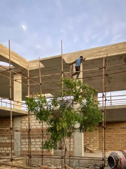 The concrete frame of a phase two building at Kizikula is completed. A worker walks along the scaffolding against the sky, highlighting the verticality of the structure and the open-air design.