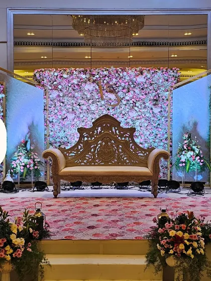 This reception stage features a dense wall of pink and white flowers, creating a stunning floral backdrop. The intricately carved traditional sofa and flanking LED screens add a blend of classic and modern elements.