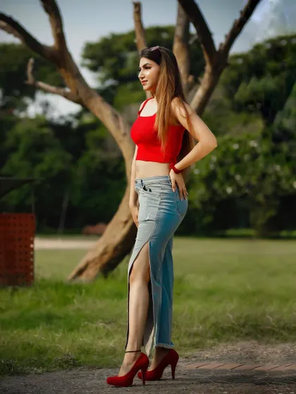 An outdoor park shoot featuring a model in a red crop top and slit jeans. The wide shot captures the full outfit and the green surroundings.
