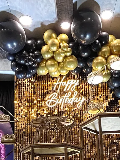 A chic and glamorous birthday celebration featuring a gold shimmer wall that catches the light beautifully. The setup is completed with a custom neon "Happy Birthday" sign and a dramatic black and gold balloon garland.
