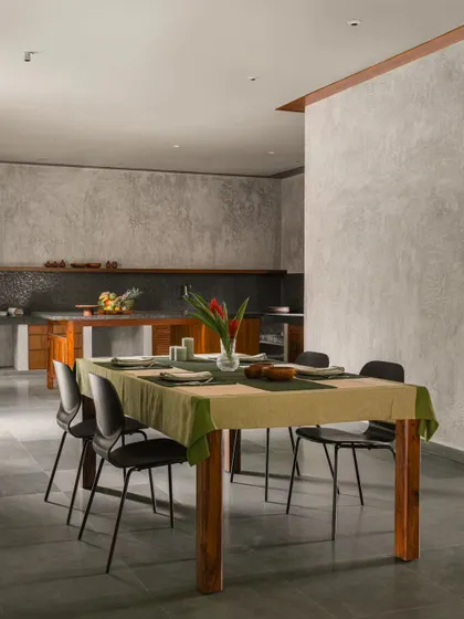 The open-plan dining and kitchen area is defined by minimalist design and functional surfaces. A simple wooden dining table is set against a backdrop of textured cement walls and a sleek, modern kitchen.
