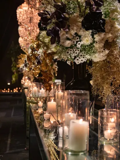 A closer view of the mirrored tablescape, showing the detail in the floral arrangements and the warm glow of dozens of candles. The composition includes rare black flowers, white blooms, and shimmering gold accents.