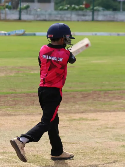 A young batter walks to the crease, bat in hand, ready to take on the opposition bowlers in a competitive match.