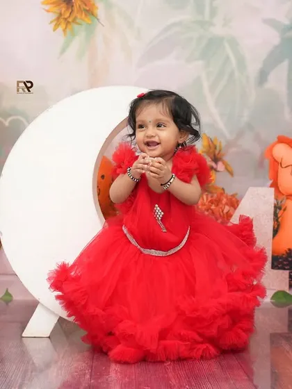 A little girl in a stunning red dress smiles brightly in a jungle-themed setup with a crescent moon prop. The mix of fairytale and nature elements creates a unique and whimsical portrait.