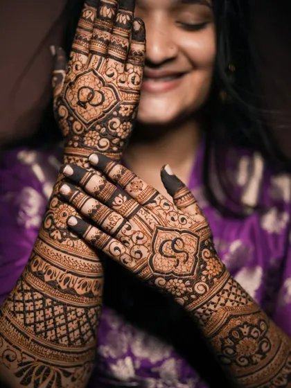 A smiling client posing with her engagement mehndi, which features her initials and ring symbols.