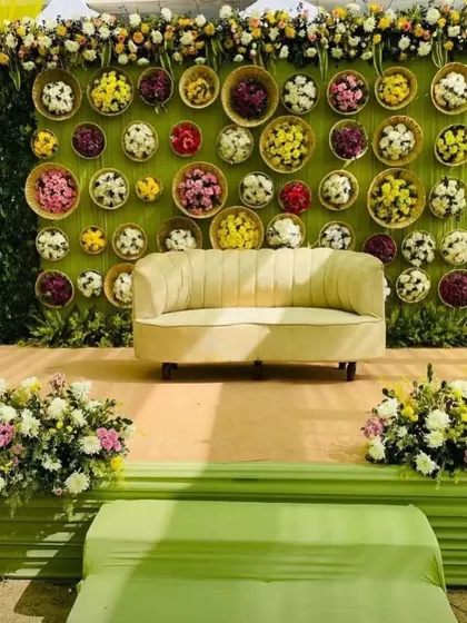 A creative and colorful stage backdrop for a Mehendi or Haldi function. The wall features small baskets filled with multi-colored flowers, creating a unique and charming photo opportunity.
