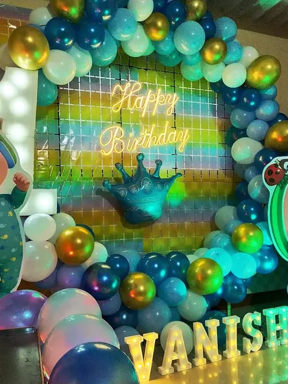 A first birthday celebration with a Cocomelon theme, using blue and green balloons, character cutouts, and a neon "Happy Birthday" sign.
