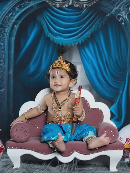 A regal portrait of a baby boy dressed as Lord Krishna, sitting on a throne with his flute.