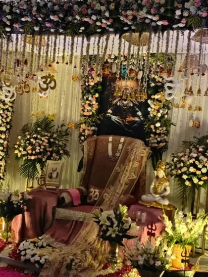 An elaborate gaddi setup for a darbar. The throne is decorated with rich fabrics and Om symbols, with a portrait of Lord Shiva in the background and flanked by large bouquets of fresh flowers.
