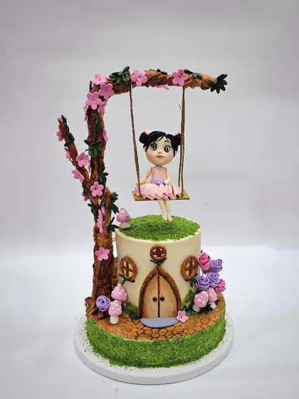 A full view of the fairytale cake, featuring a fairy on a swing under a blossoming tree, all part of a gravity-defying structure. The base is a hobbit-style house with a detailed wooden door and tiny mushrooms.