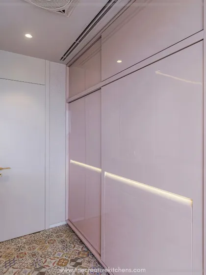 A full view of the glossy pink PU sliding wardrobe. The inbuilt shutter lights cast a mesmerizing glow, illuminating the narrow passage and making the space feel larger and more luxurious.