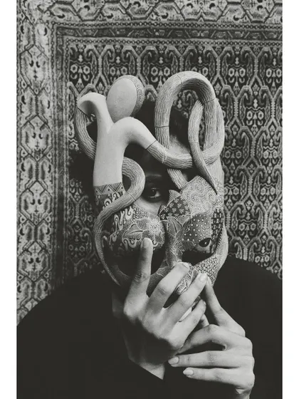The same concept as the previous shot, rendered in dramatic black and white. The monochrome treatment emphasizes the texture of the mask and the intensity of the model's visible eye.