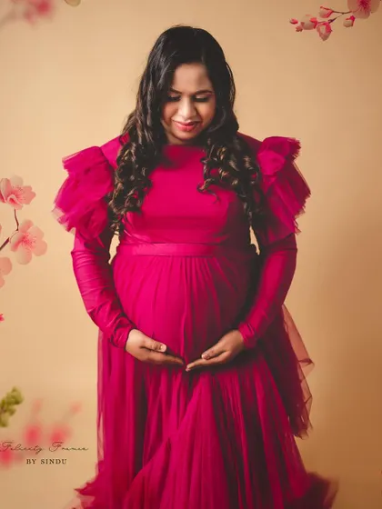 A close-up of an expectant mother in a beautiful magenta tulle gown, gently holding her belly. The soft lighting and floral accents create a feeling of warmth and new beginnings.