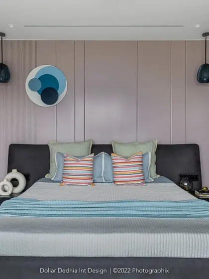 A full view of the pastel bedroom, with its lavender paneled wall, dark grey upholstered bed, and black accent lights that ground the soft color scheme.