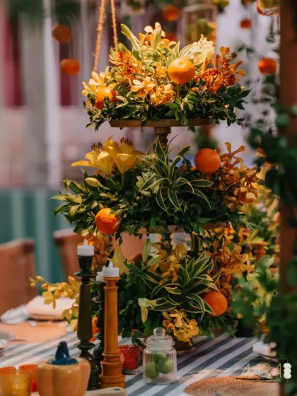 A stunning three-tiered floral centerpiece from our 'Borough Bazaar' theme. The arrangement combines vibrant orange lilies and orchids with fresh oranges, creating a lush and aromatic focal point for the table.