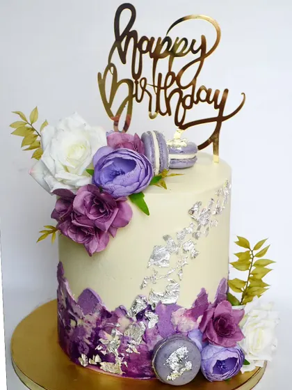 A gorgeous cake in shades of purple. The design includes abstract buttercream textures, silver leaf, macarons, and a beautiful arrangement of purple flowers.
