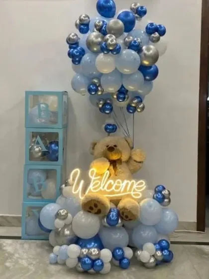 A welcome decoration for a baby boy. This setup features a large teddy bear holding a 'Welcome' neon sign, surrounded by balloons in shades of blue and silver, and complemented by 'BABY' display boxes.