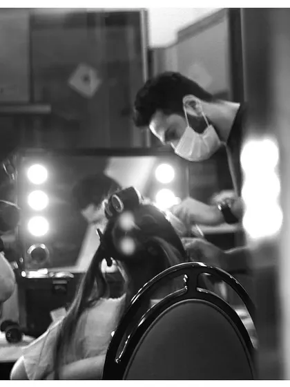 A black and white shot of our hairstylist at work, focused on creating the perfect look in front of a professional lighted mirror. This captures the focused, behind-the-scenes atmosphere.