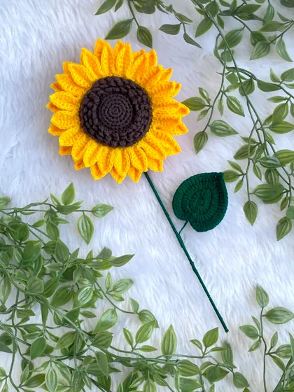 A beautiful, large crochet sunflower with a single leaf, lying on a soft white background. The vibrant yellow and rich brown center are so eye-catching.